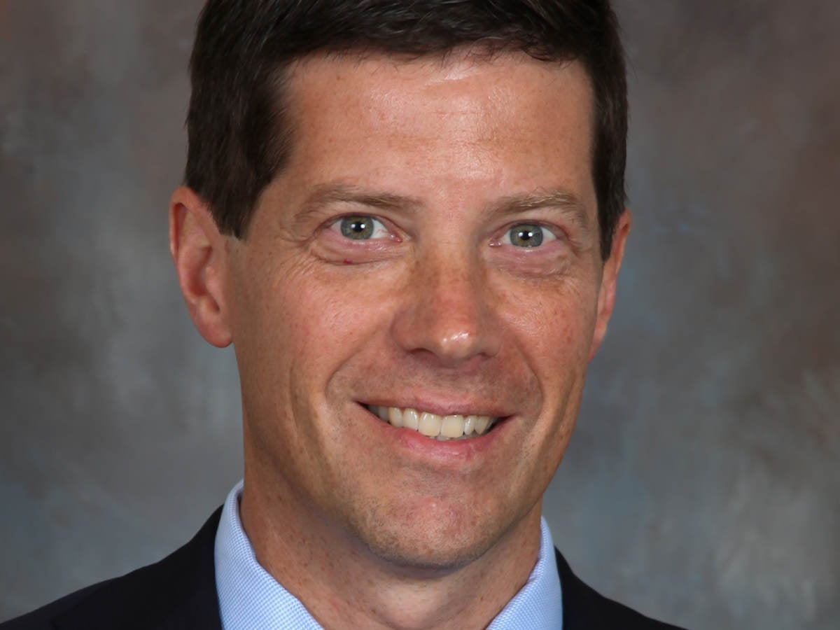Headshot of Todd Denison wearing a black suit, blue shirt and navy blue tie on a grey background