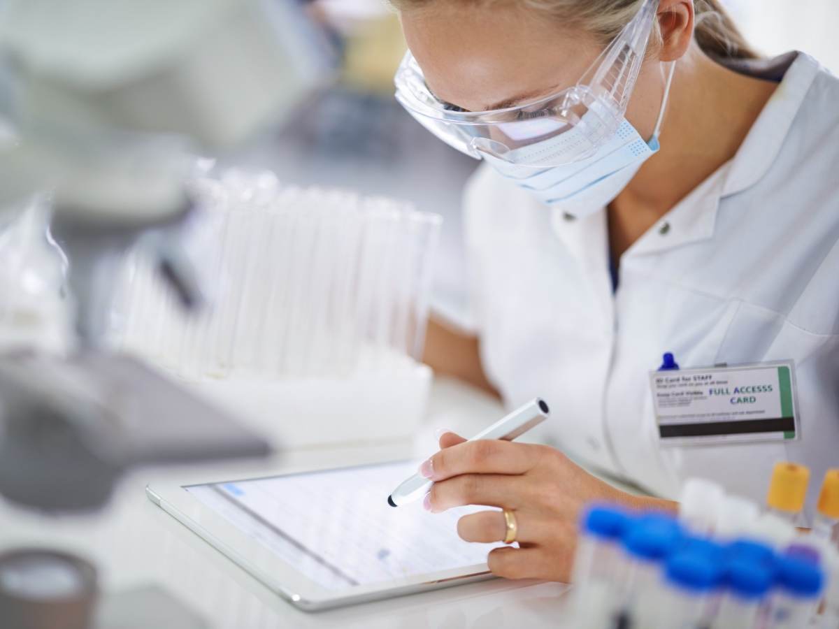 Female medical professional wearing protective eyewear and facemask making notes on electronic device