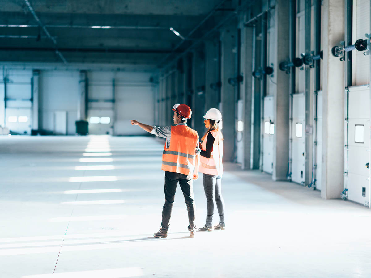 Two people in a warehouse wearing construction hats and vests pointing and looking at something
