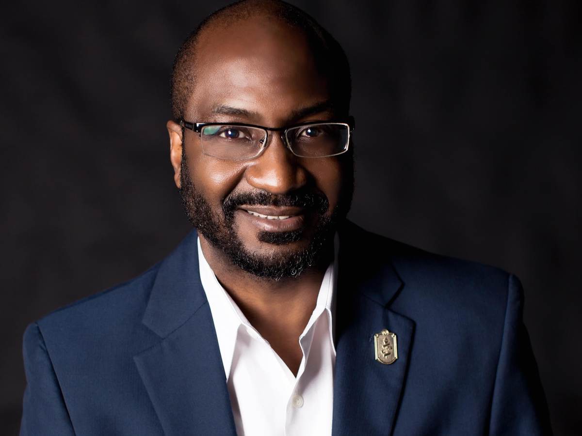 Dwayne Sloan poses for headshot wearing a navy blue jacket, white collar, unbuttoned shirt on a dark background.