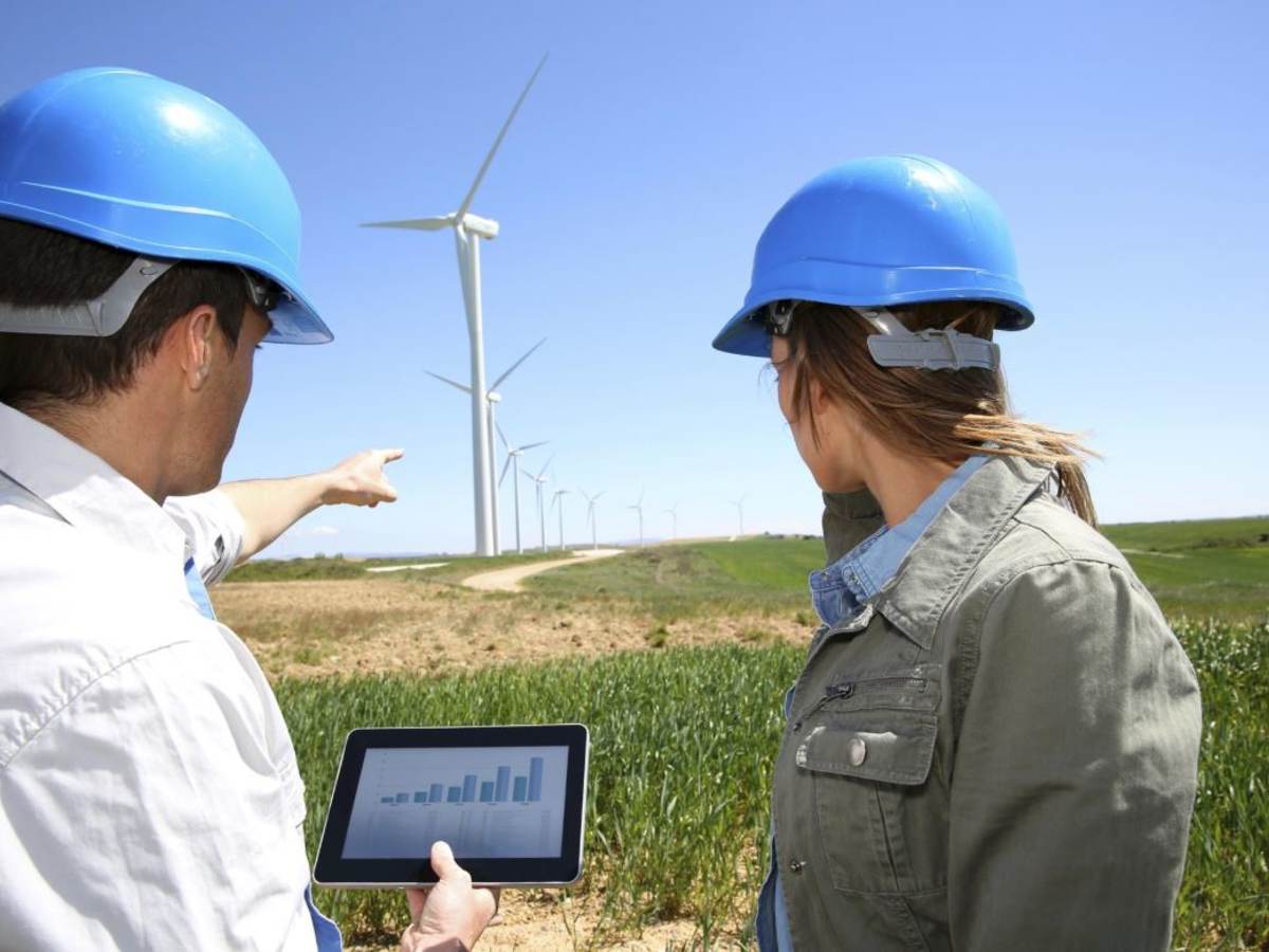 Technicians in blue hard hats standing in a field with an iPad while pointing at wind turbines