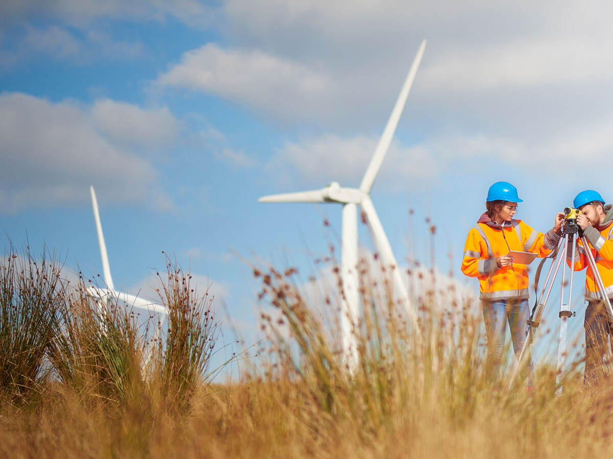 Two workers surveying in a field with wind turbines behind them