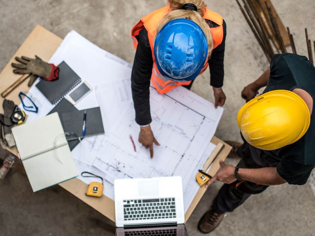 Two construction workers examine blueprints with laptop open on a project table