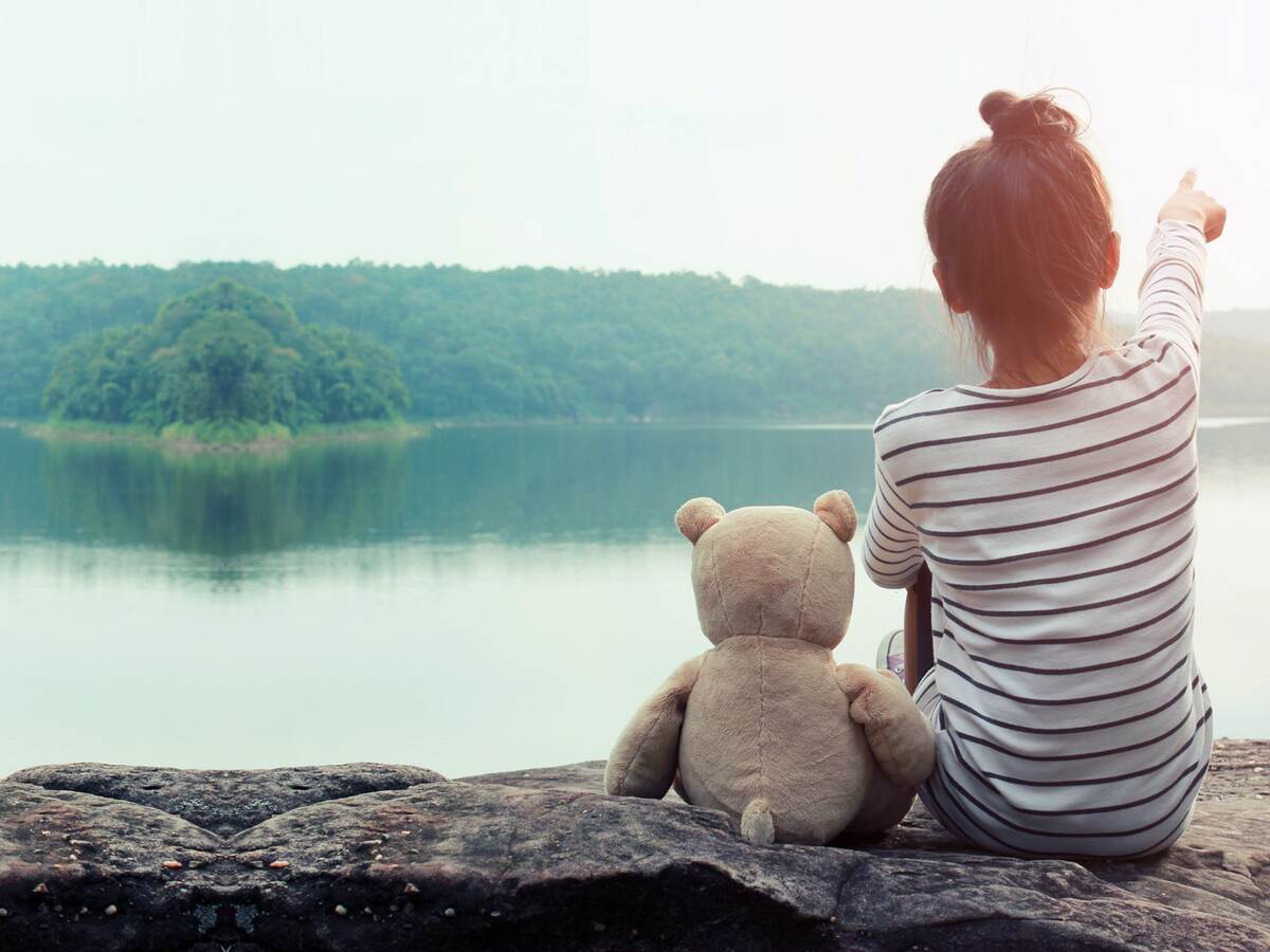 A girl sitting at a lake with a stuffed teddy bear next to her