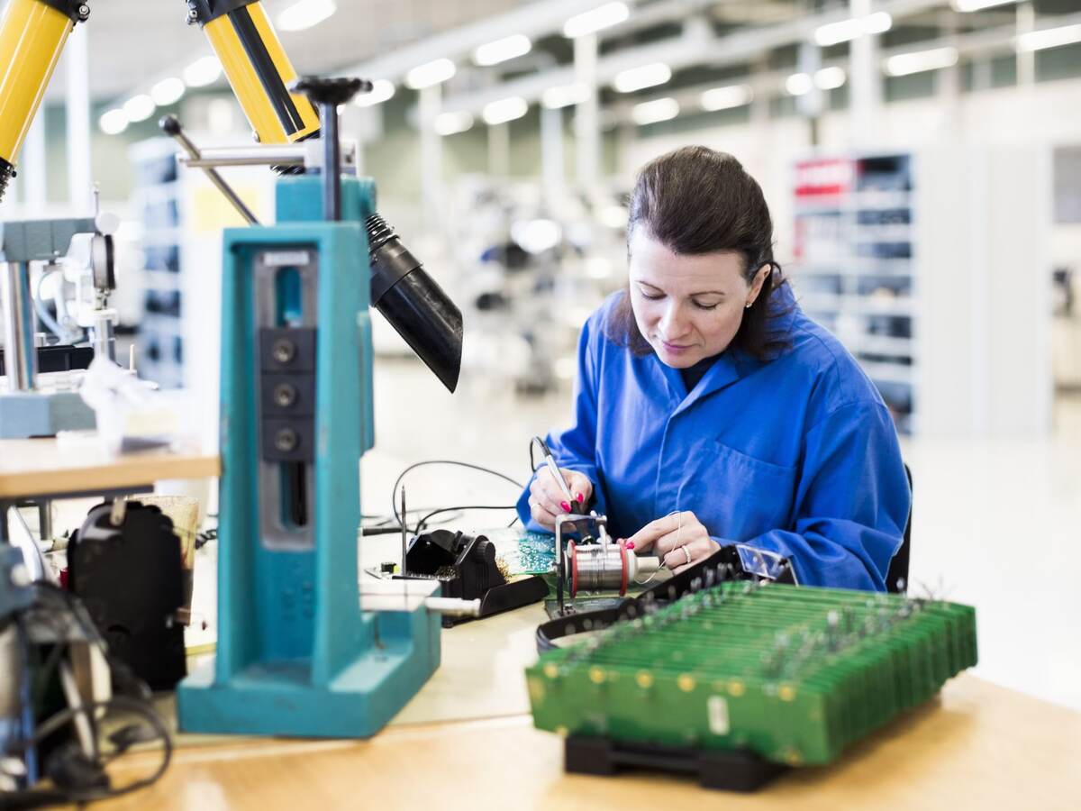 Mid adult female electrician working on circuit board at desk in industry.