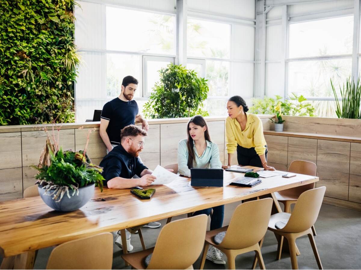 In a modern office with a green plant wall and windows, a group of young co-workers collaborate around a modern wooden dining table.